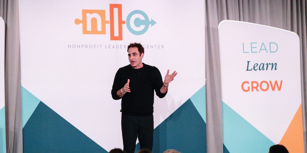 Matthew Emerzian a white man with brown hair wearing a black shirt and pants stands on the stage at the Nonprofit Leadership Conference speaking to 700 nonprofit leaders about the importance of mattering