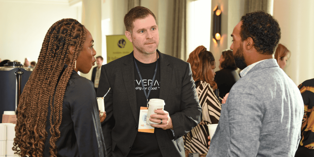 Jay Taylor, CEO of Leverage, talking to two NLC board members. Jay is a white male with brown hair wearing a Leverage T-shirt under a black jacket