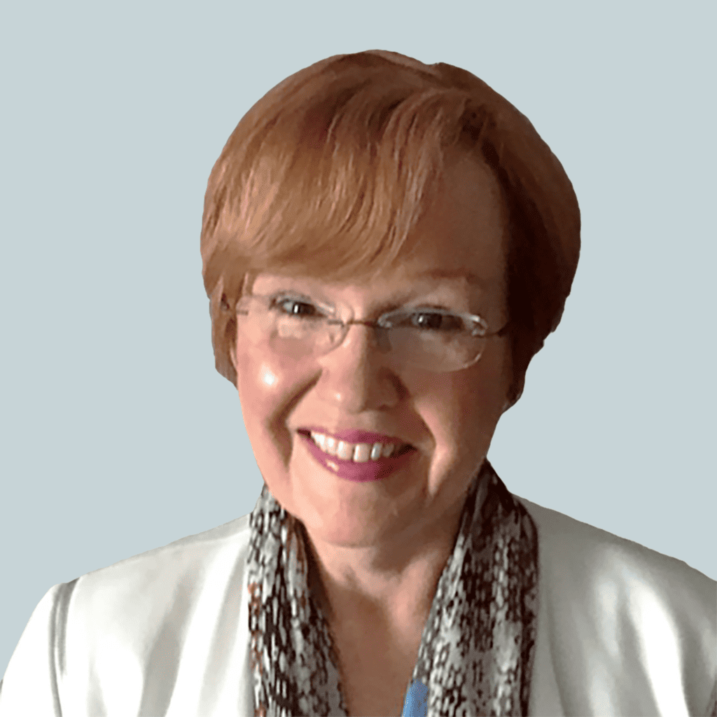 Headshot of Abby Mackness, a white woman with short, reddish-brown hair, wearing glasses, a white jacket and floral scarff