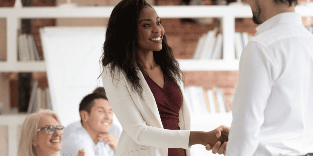 A Black woman wearing a white jacket and maroon top shakes the hand of a white man in a white button-down shirt
