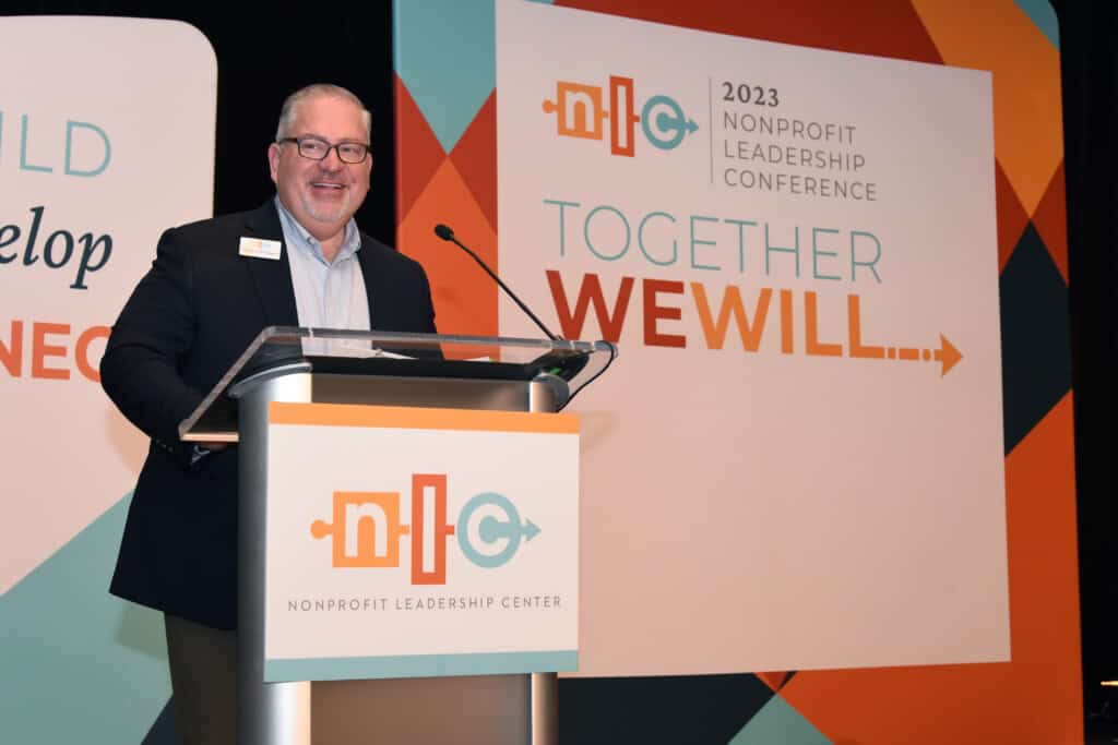 Charlie Imbergamo, MA, CFRE, a white man with greying hair and glasses wearing a dark grey suit and standing on a podium with the NLC logo on it