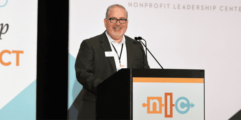 Charlie Imbergamo, the CEO of the Nonprofit Leadership Center speaking at the podium; a white male wearing glasses and a grey suit