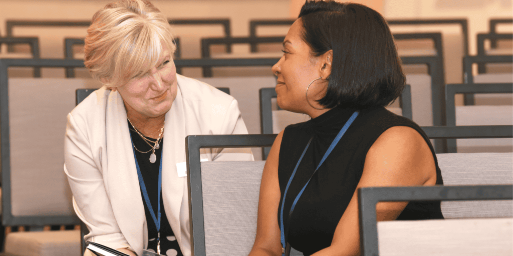A white woman with blonde hair wearing a white jacket sits talking to a Black woman with dark brown hair and a black top