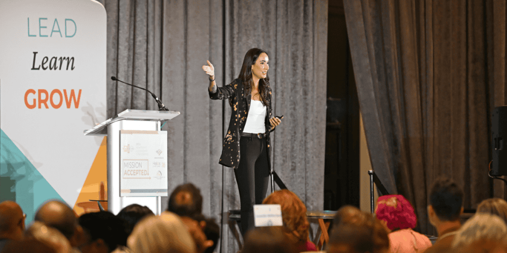 Sam Rad a white woman with long brown hair wearing a black suit speaks on stage at the Nonprofit Leadership Conference