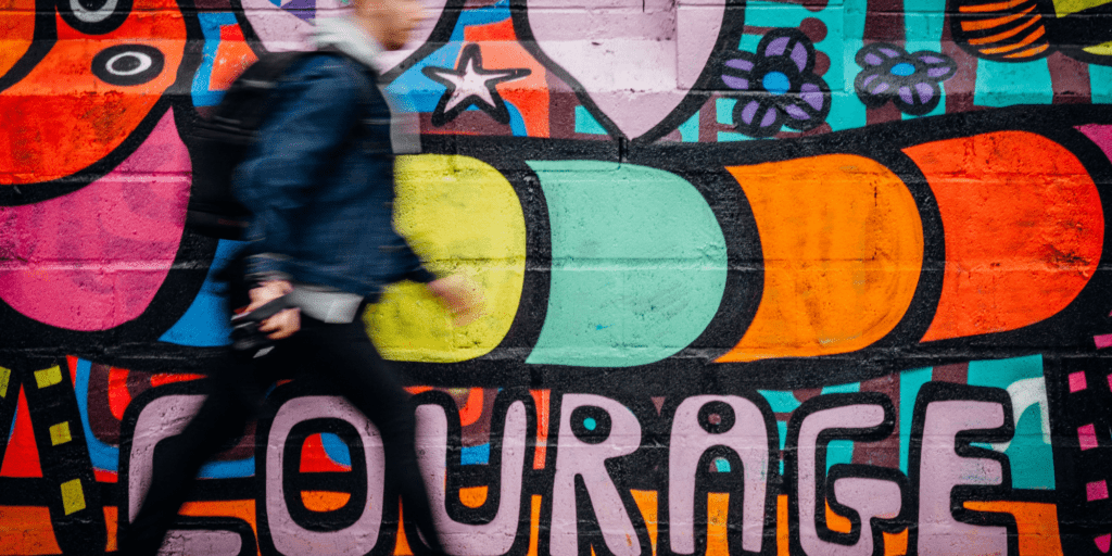 White man walking, blurred, against a painted background that says courage: yellow, blue, orange red and purple in the mural