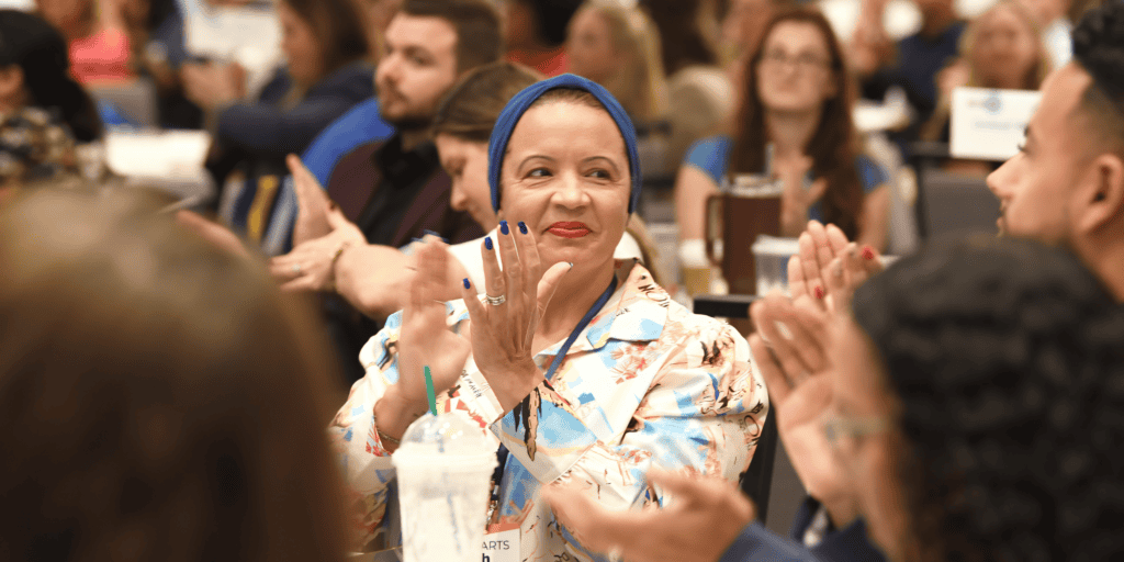 A Black woman wearing a blue headband and floral top is clapping in a crowd of other leaders