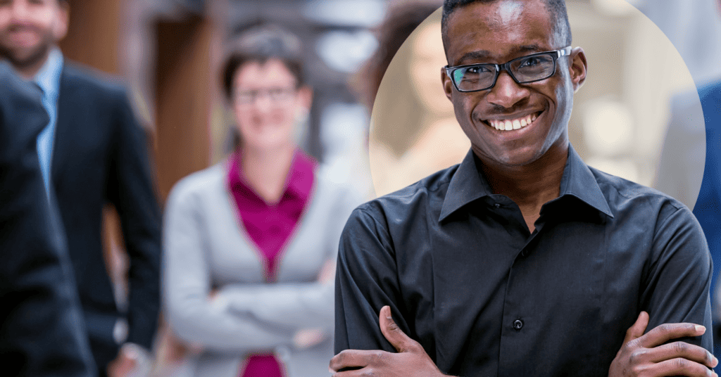 A Black man in his 30s wearing a black shirt and smiling at the camera with blurred colleagues in the background