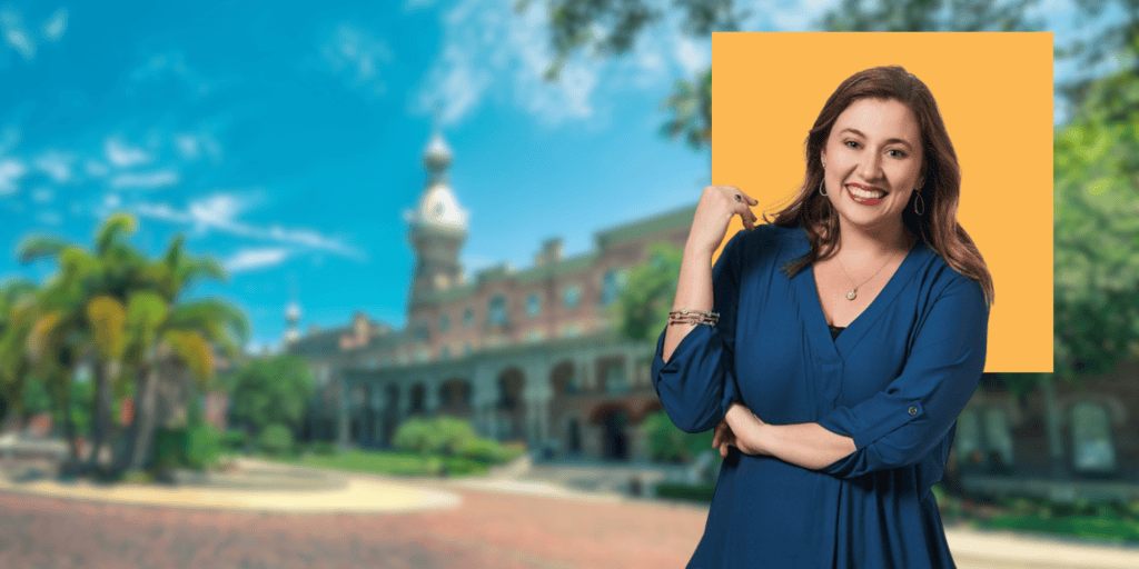Photo of Jessica Dvoracsek, a white woman with dark brown hair wearing a royal blue top smiling into the camera. Standing in front of a scene of the University of Tampa.
