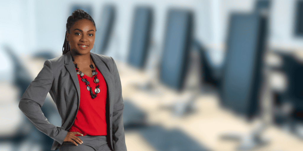 A photo of Kadian Douglas, a Black woman of Dominican descent wearing a grey suit and red blouse, featured in front of a blurred background of computers at a desk