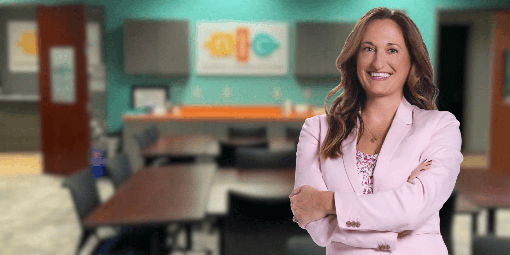 Photo of Katie Shultz, a white woman with long brown hair wearing a pink suit jacket smiling at the camera with the NLC training center blurred in the background