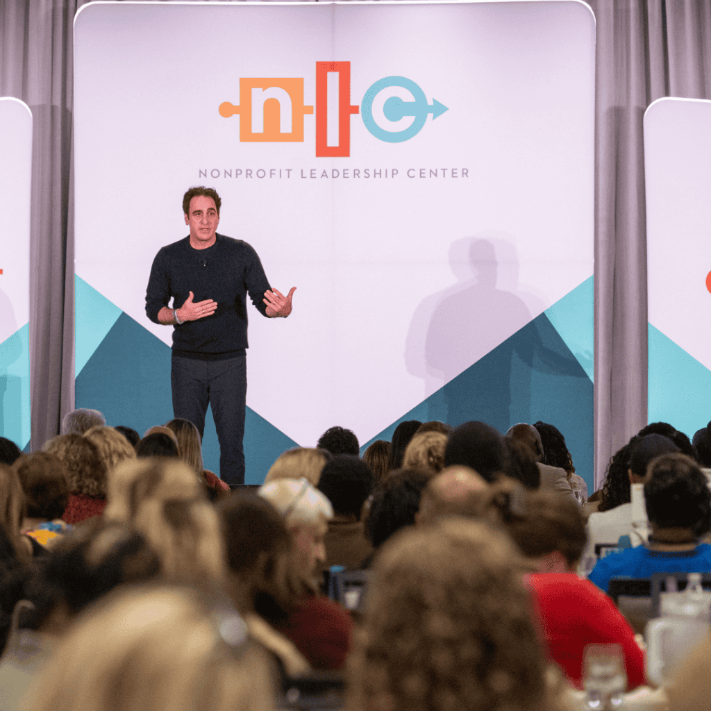 Matt 3 Nonprofit Leadership Center of Tampa Bay Male speaker stands on stage gesturing with hands addressing a seated audience at a nonprofit leadership conference with a large banner that reads Nonprofit Leadership Center in the background