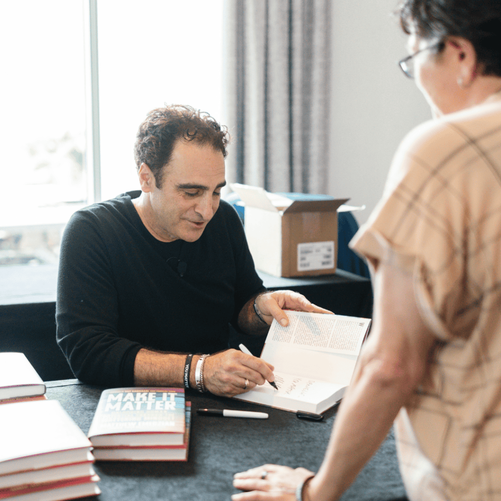 Matt 4 Nonprofit Leadership Center of Tampa Bay Author signs a book at a table during a book signing with stacks of copies nearby and a reader standing beside him in the foreground
