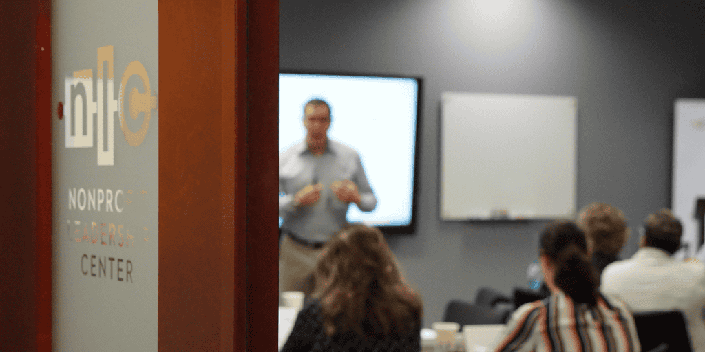 Partially open door with the words NLC Nonprofit Leadership Center with a blurred image of a class in session with nonprofit professionals