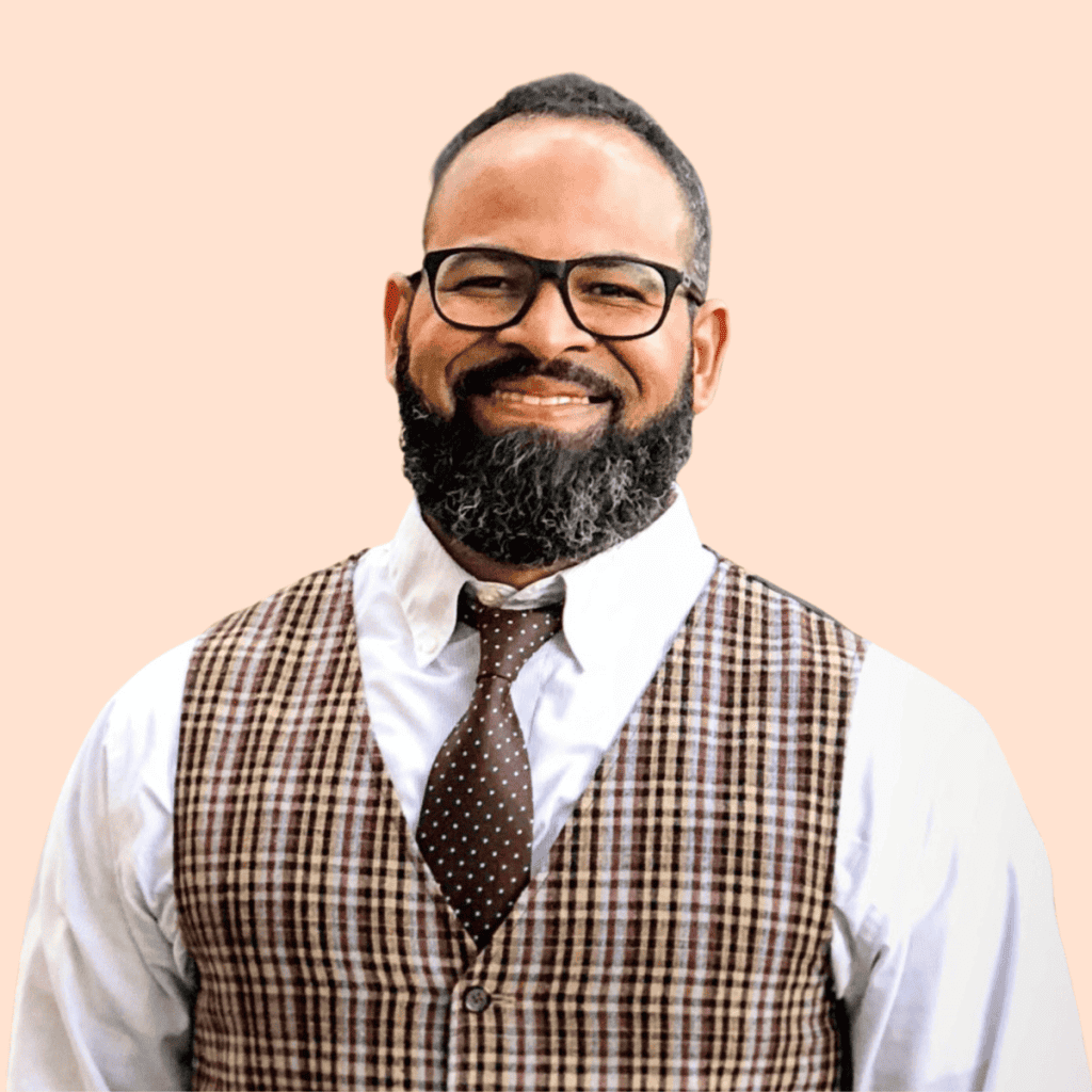 Nestor Ortiz, a Black man with dark hair and a beard, wearing glasses and a white button-down shirt and plaid vest, smiling into the camera