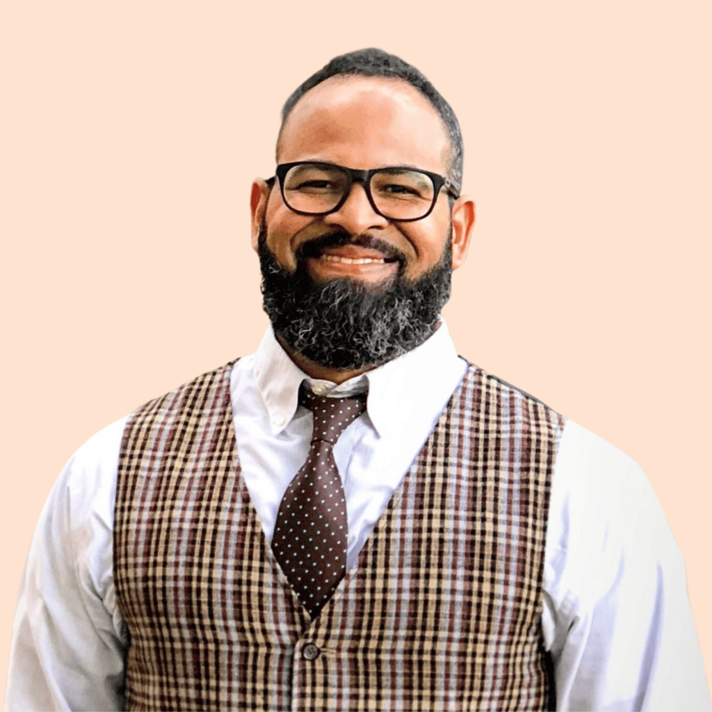 Nestor Ortiz, a Black man with black hair and a black beard wearing glasses, a white shirt and brown tie and vest against a peach background