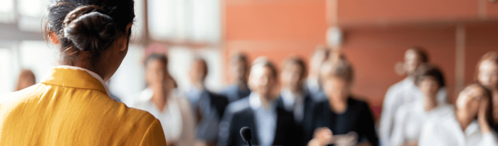 Photo of a Black woman from the back speaking to a crowd of diverse professionals who are slightly blurred