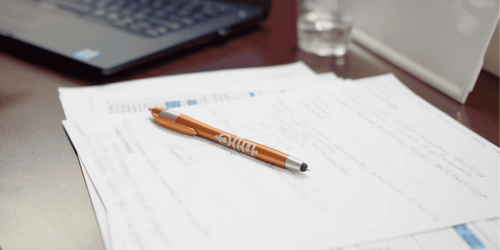 Orange NLC-branded pen sitting on a stack of notes on a desk