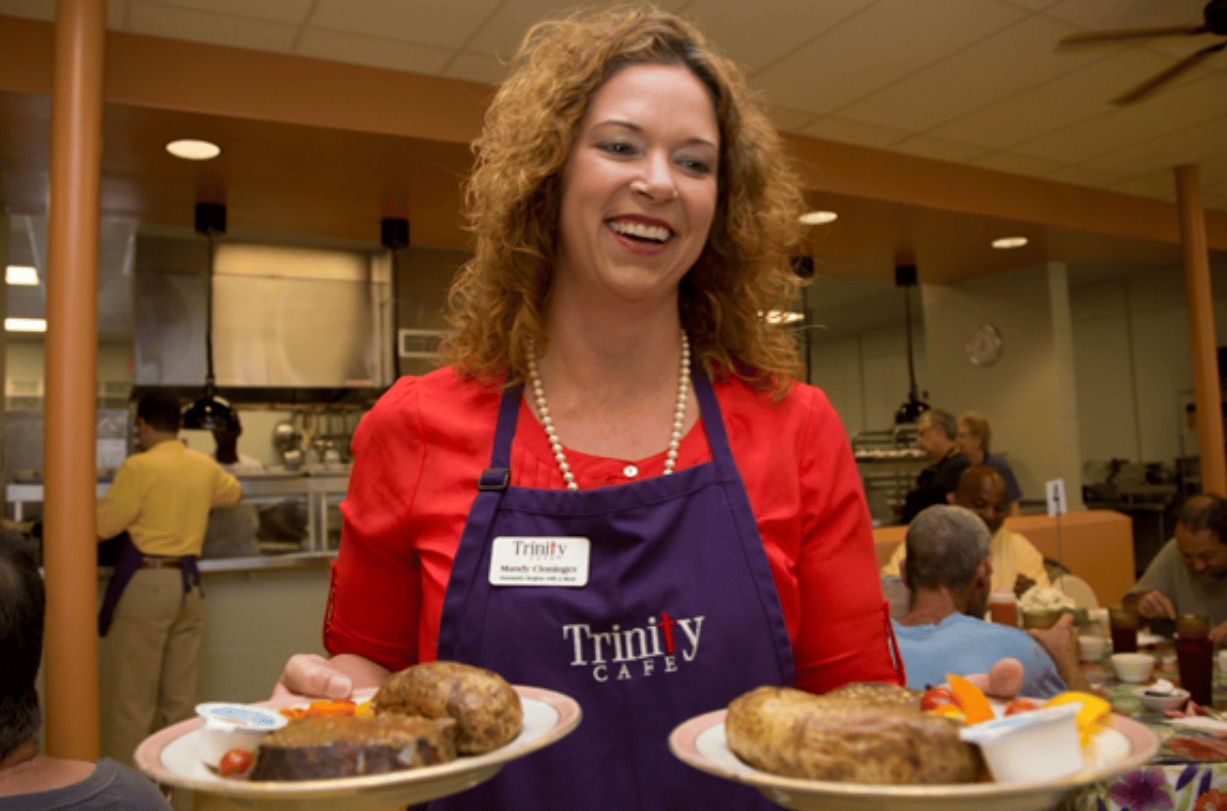 Mandy Cloninger holds two plates of hot food that she carries to a table. She is wearing a red top and a blue apron that says Trinity Cafe