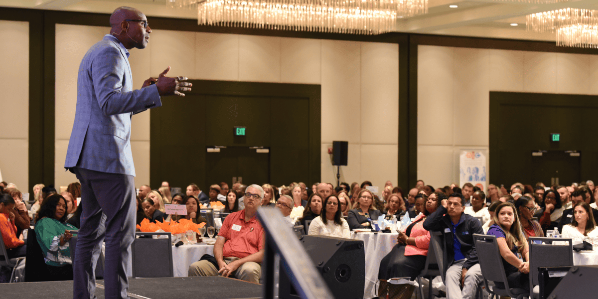 Shola Richards, a Black man and best-selling author speaks on stage in a grey suit to a large, diverse group of nonprofit professionals