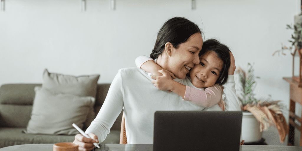 An Asian woman with dark hair and a white top working at her laptop while hugging her daughter
