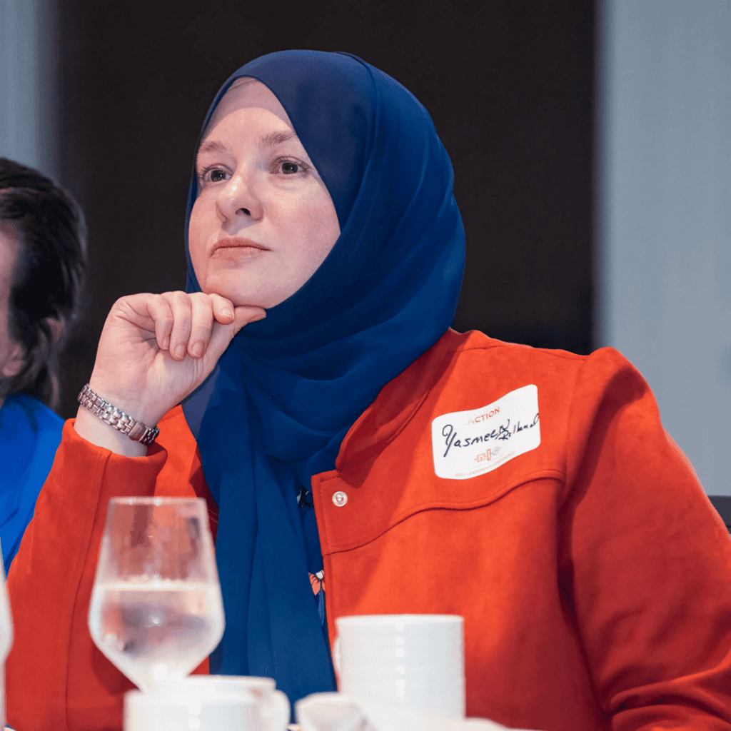 Yasmeen Rolland, a middle-Eastern woman wearing a red jacket and blue head covering listening to a speaker