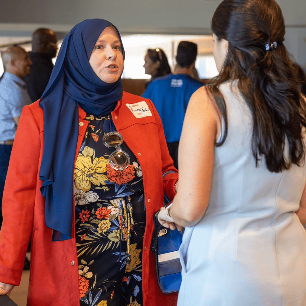 Yasmeen Rolland, a middle-Eastern woman wearing a red jacket and blue head covering speaking to another woman in a light blue dress with dark brown hair