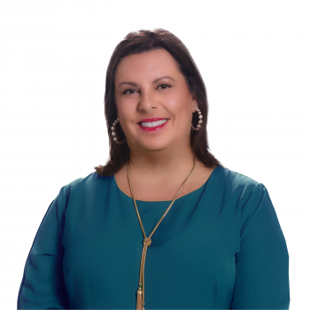 A white woman with shoulder-length brown hair wearing a blue top and gold necklace