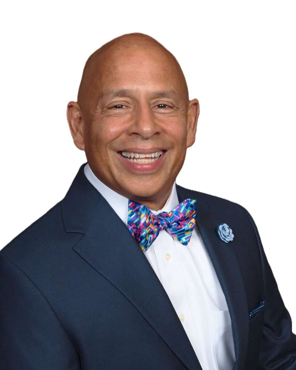 Smiling man wearing a navy suit white shirt and a bright multicolored bow tie cropped to head and shoulders